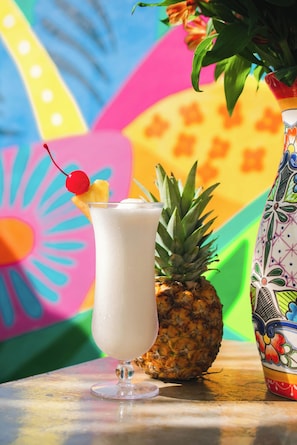 Un vaso de una bebida cremosa con cereza en el borde, una piña y un jarrón colorido sobre una mesa.