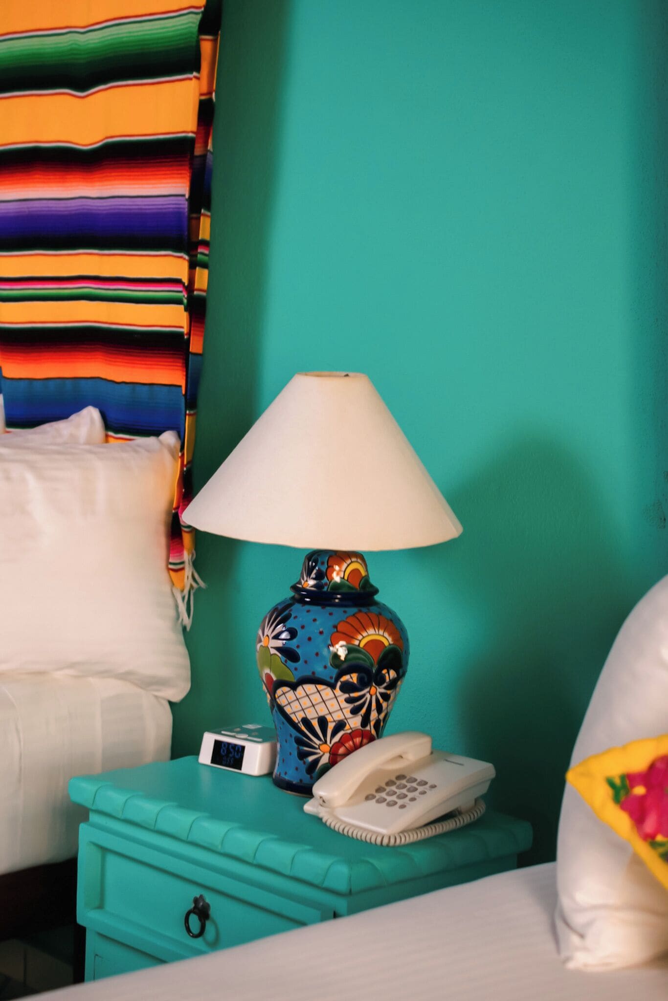 Una mesita de noche azul turquesa con una lámpara estampada, un almohadón blanco y un pequeño reloj digital.