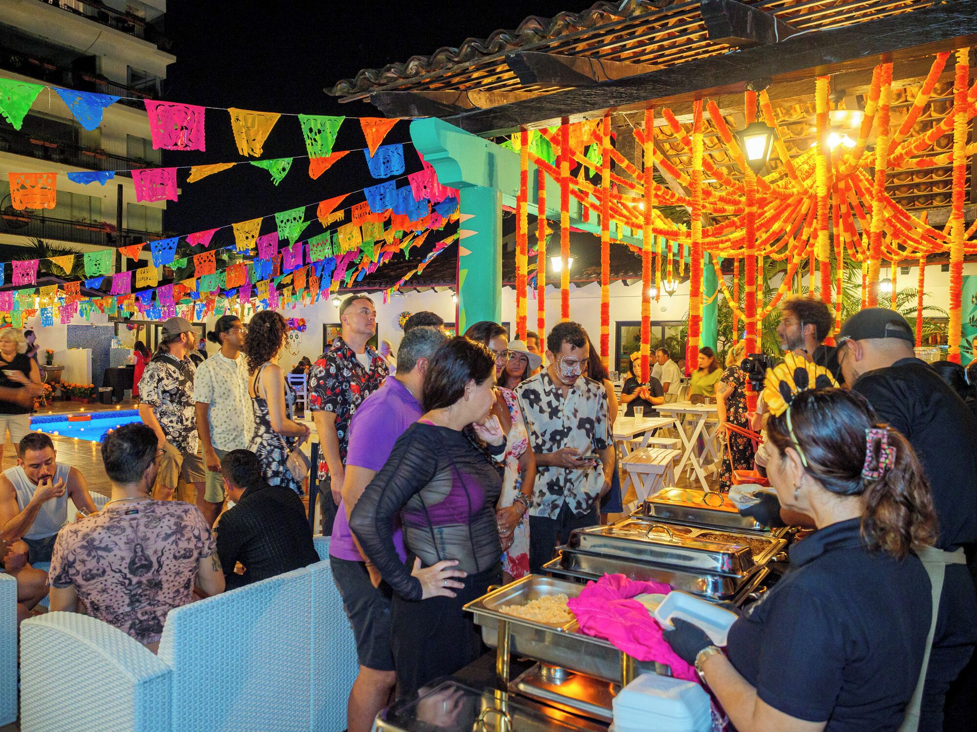 A crowded outdoor event with people gathered around food service areas, decorated with colorful paper flags and floral arrangements.