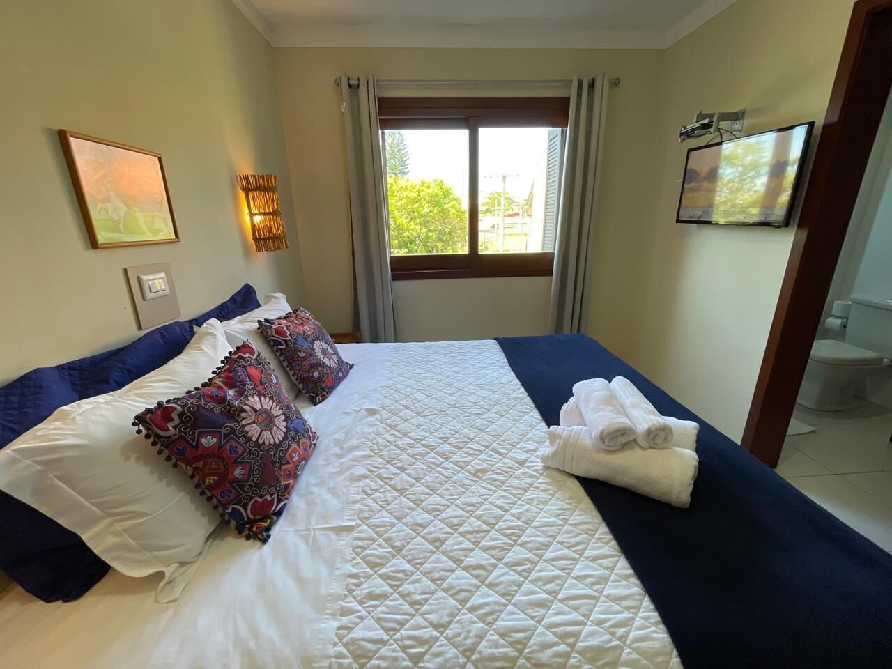 Una cama bien hecha con un cubrecolchón acolchado, dos almohadas decorativas y un juego de toallas sobre un corredor azul.