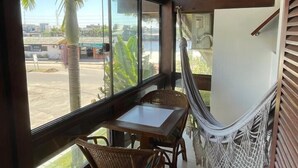 Una habitación con techo de madera, una mesa con sillas, una hamaca y ventanas amplias que dan vistas a palmeras y edificios.