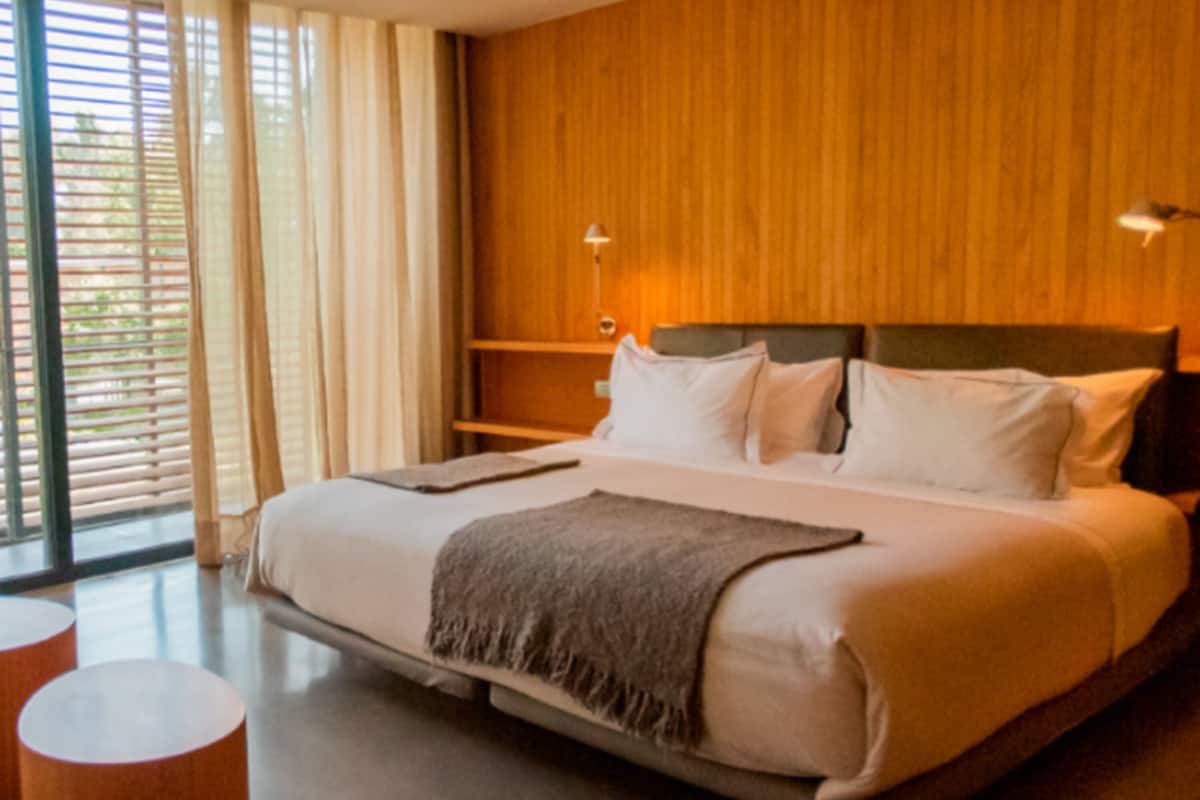 A modern hotel room with a large bed, wooden paneling on the walls, a bedside table with a lamp, and a view through the window.