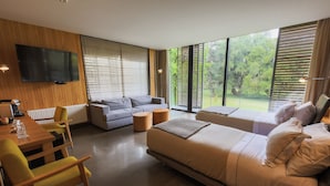 A modern hotel room with a large bed, a sofa, a dining table with chairs, and a TV mounted on the wall.