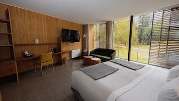 A modern hotel room with a large bed, a desk with a chair, a television, and a view of trees through the window.