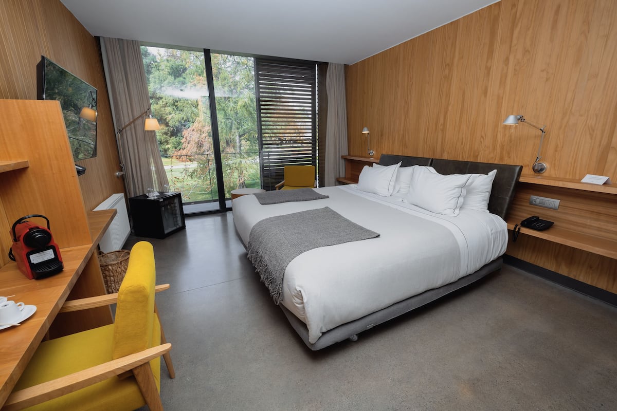 A modern hotel room with a large bed, a desk with a chair, a desk lamp, and a view of the outdoors through large windows.