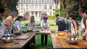 A group of people in aprons around a wooden table with metal bowls, in front of a large building with a garden.