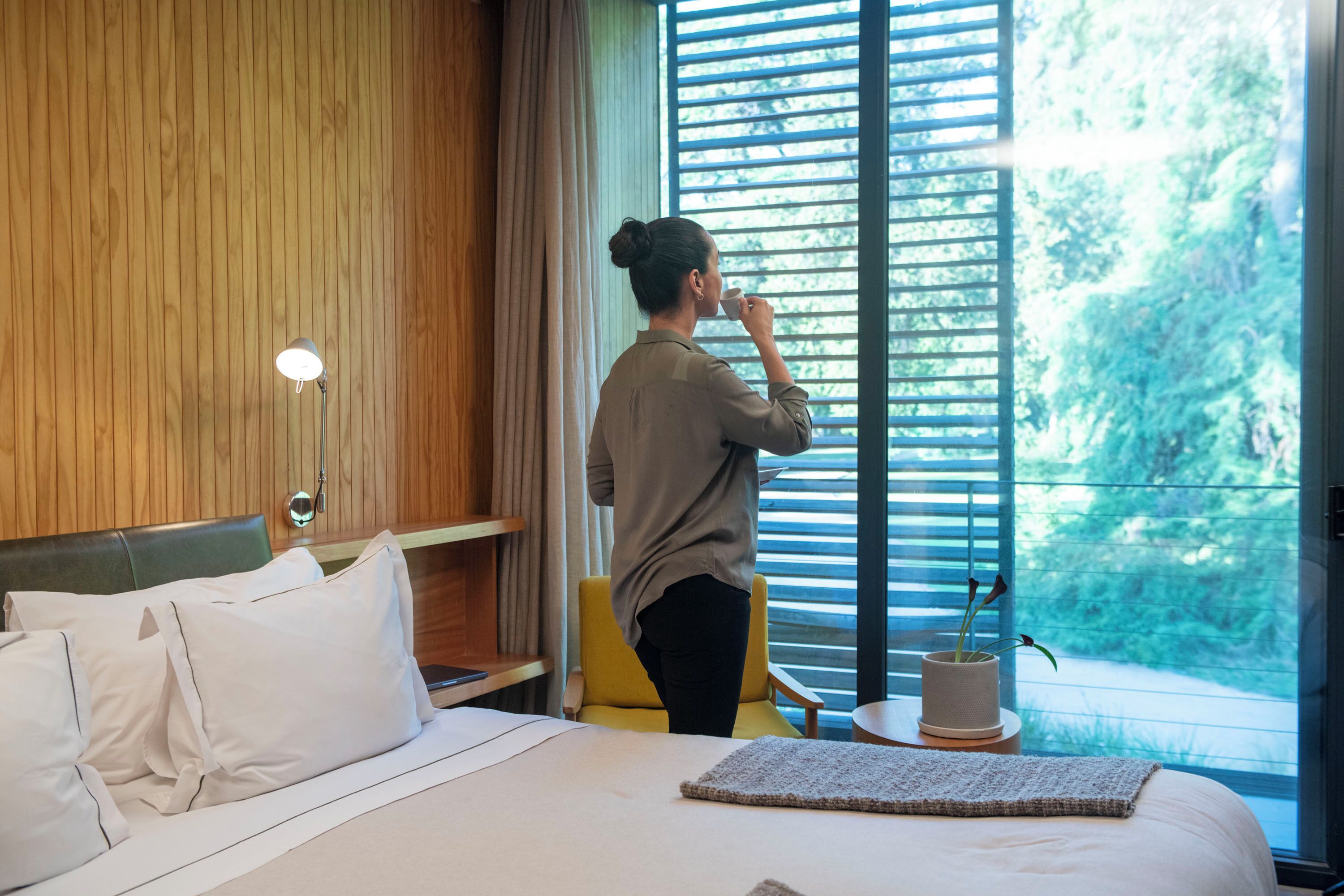 A hotel room with a bed, a bedside table, a lamp, a window with blinds, and a view of greenery outside.