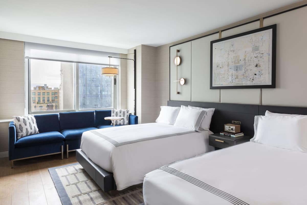 Quarto de hotel com duas camas, um sofá, uma mesinha e uma janela grande com vista para a cidade.