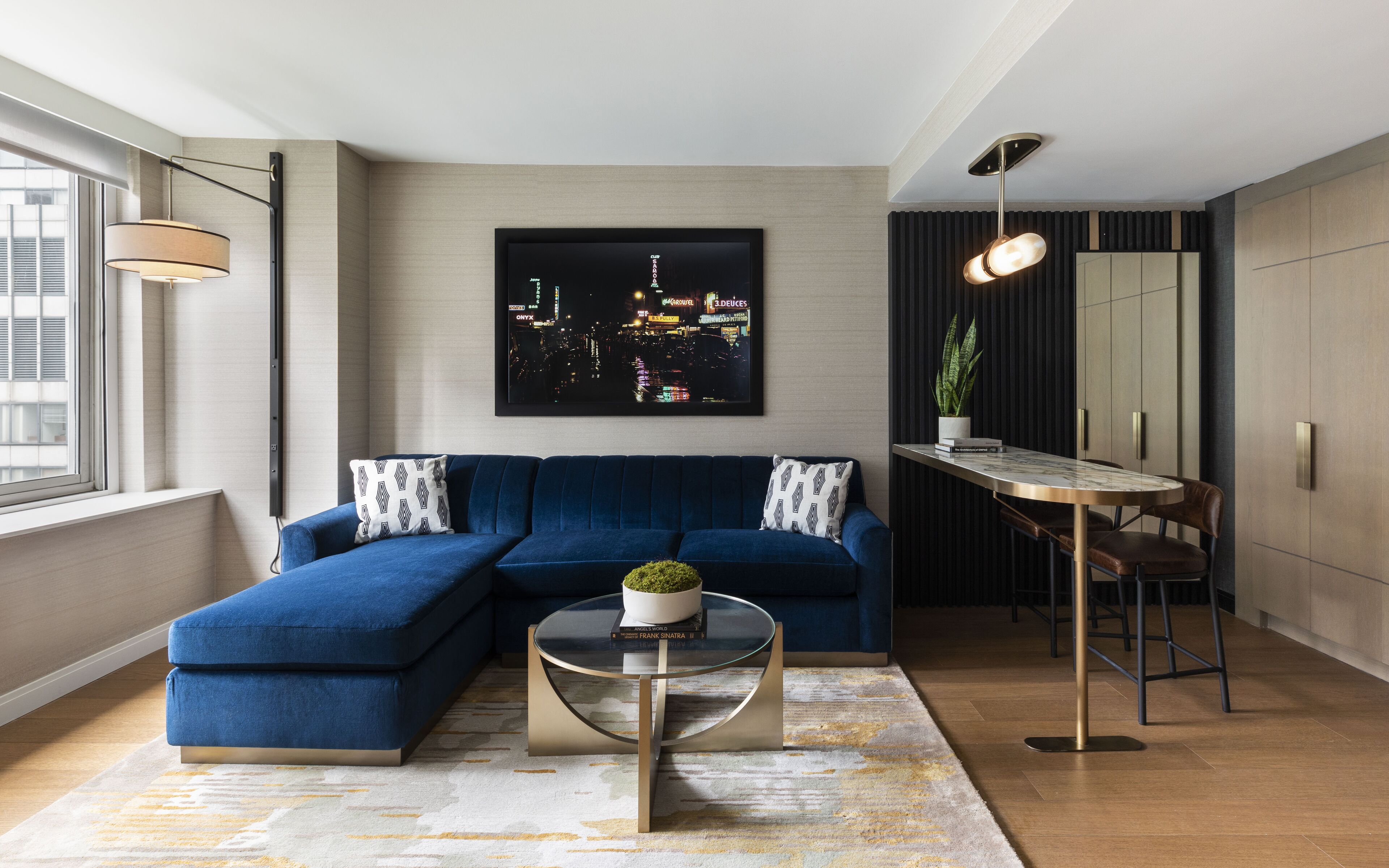 A modern living room with a blue sectional sofa, a glass coffee table, a cityscape painting, a dining area with a small table and chairs, and a large mirror.