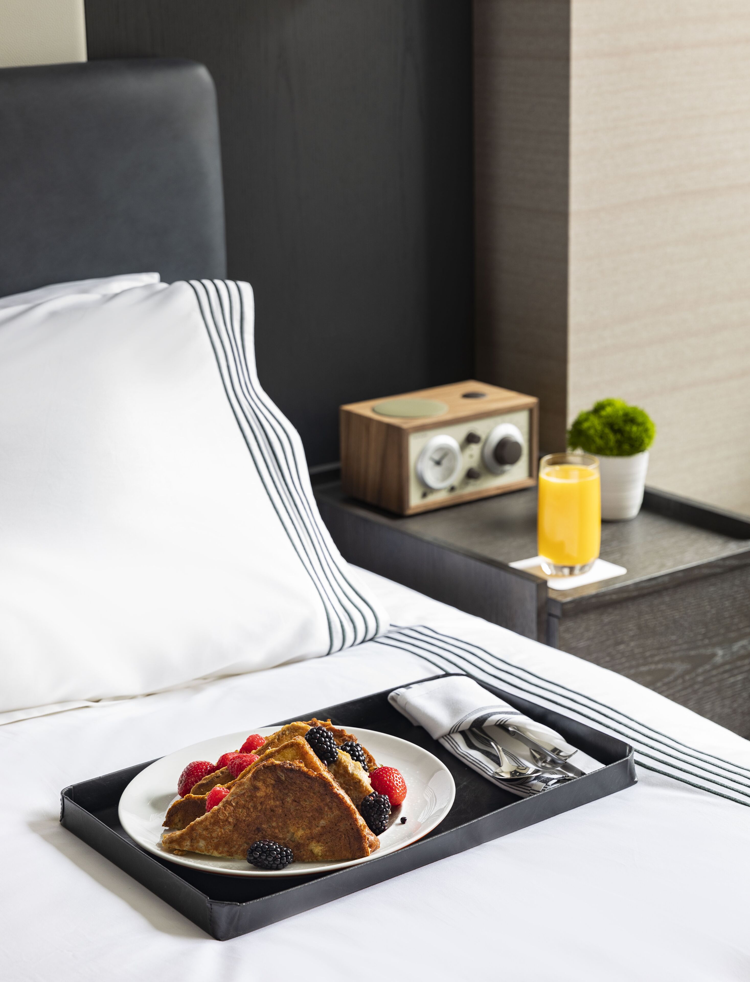 A neatly made bed with a white pillow and a black and white striped blanket. A tray with a plate of food, including a slice of cake and berries, is placed on the bed. A wooden radio and a glass of orange juice are on a side table next to the bed.