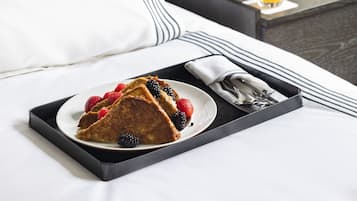 A neatly made bed with a white pillow and a black and white striped blanket. A tray with a plate of food, including a slice of cake and berries, is placed on the bed. A wooden radio and a glass of orange juice are on a side table next to the bed.