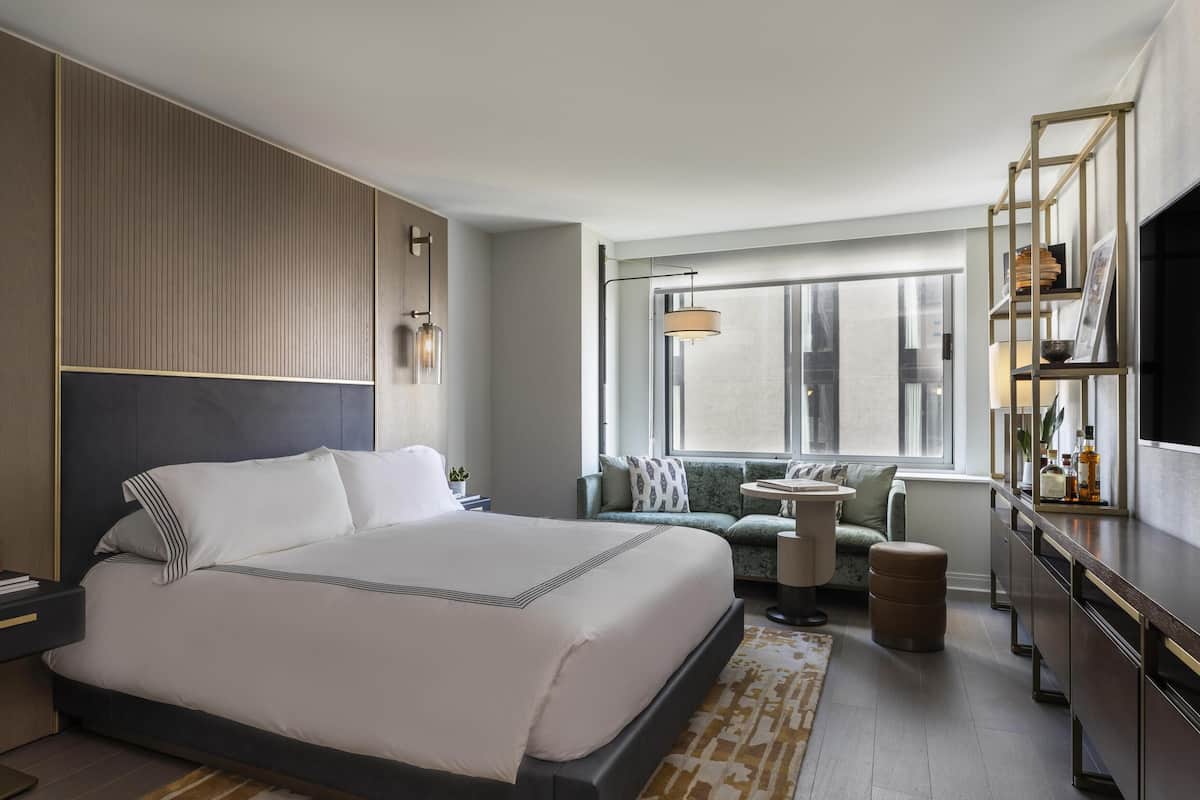 Quarto de hotel moderno com uma cama grande, área de estar, televisão e estante.