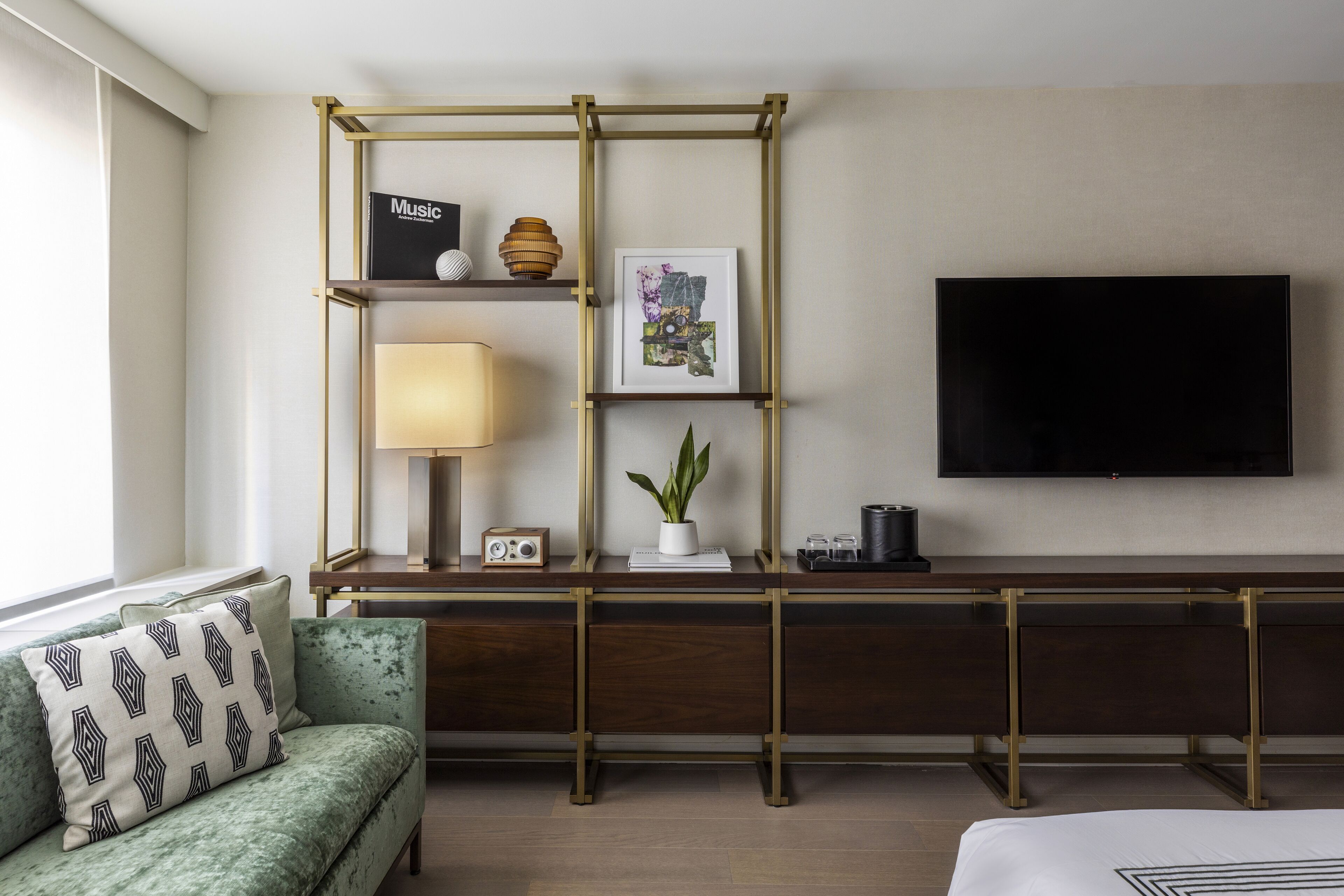 Habitación de hotel con sofá verde, un escritorio con lámpara, un televisor de pantalla plana y una estantería con varios objetos.