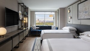 Moderni hotellihuone, jossa on kaksi sänkyä, televisio, sohva ja suuri ikkuna, josta avautuu näkymä kaupunkiin.