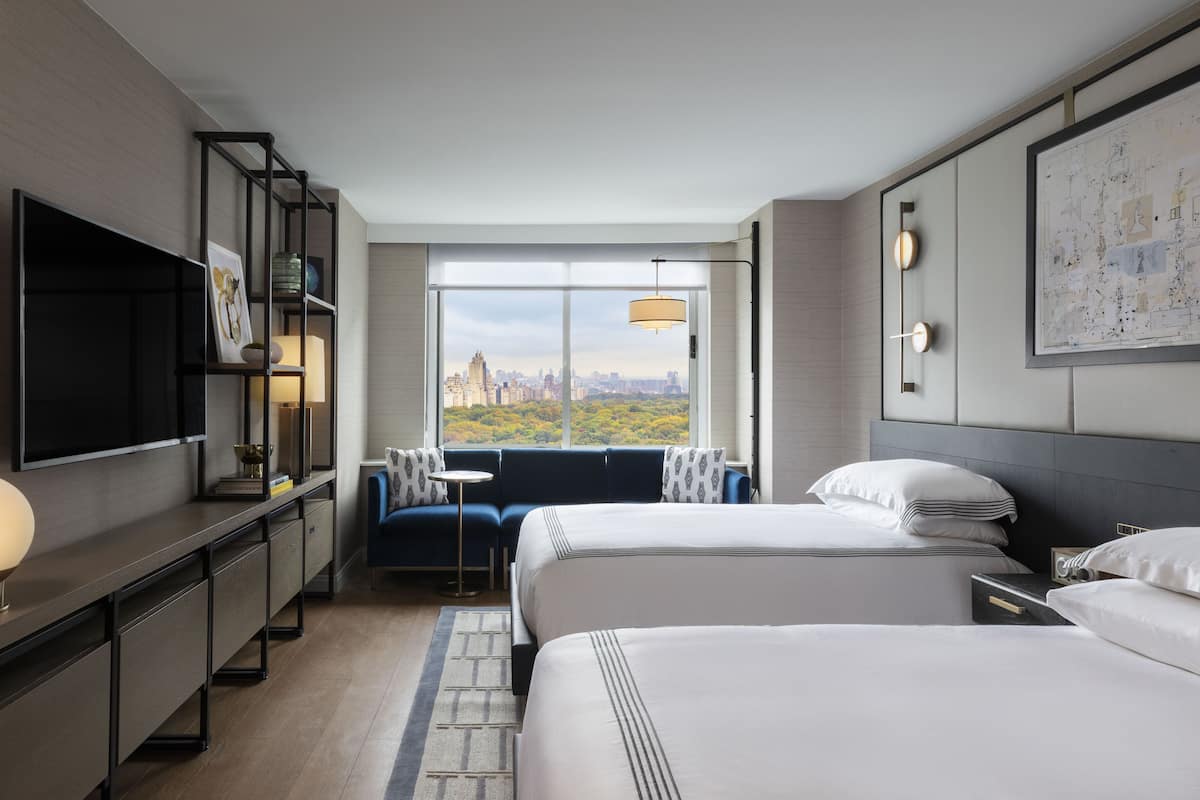 Quarto de hotel moderno com duas camas, televisão, sofá e uma janela grande com vista para a cidade.