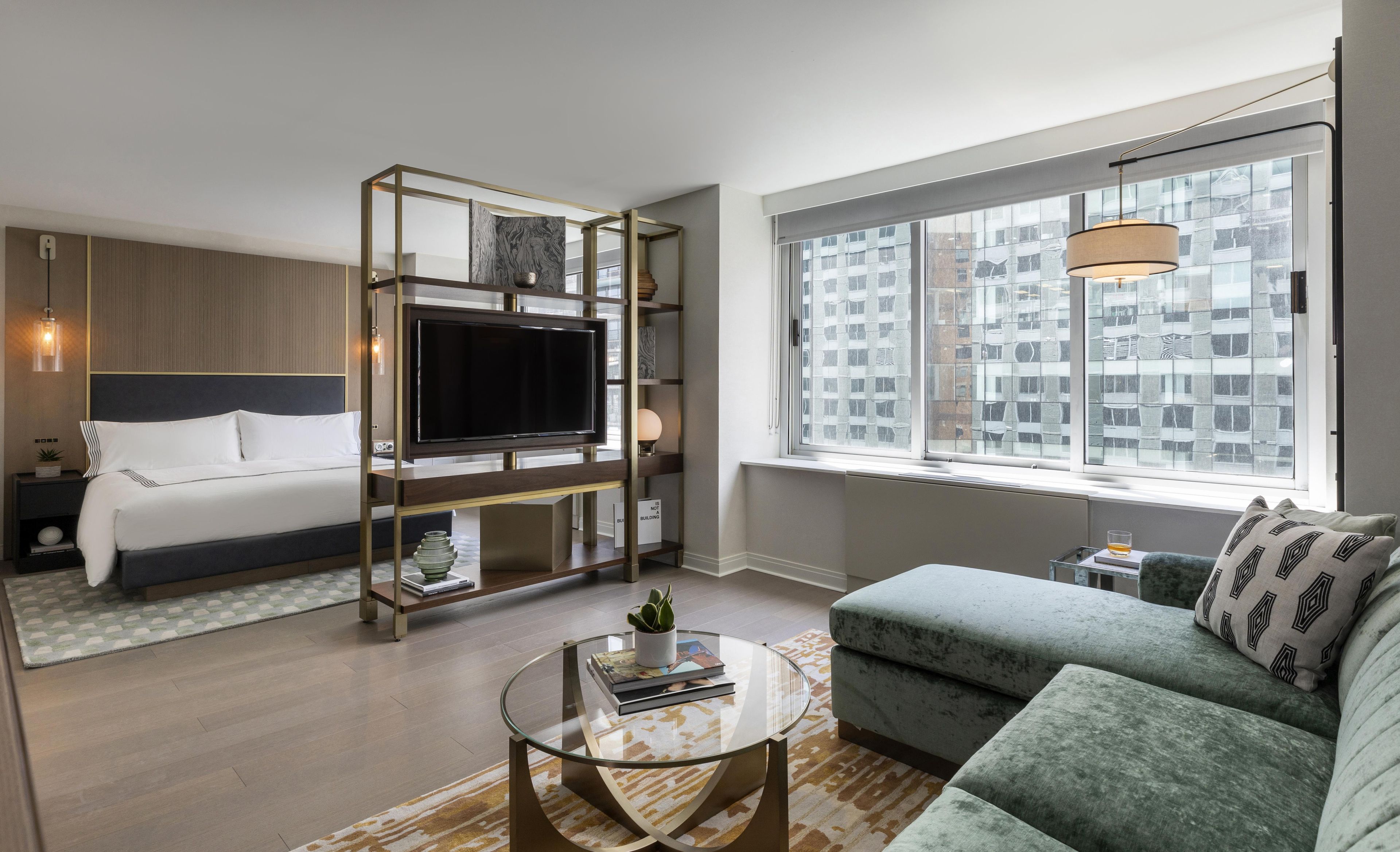A modern hotel room with a bed, a TV mounted on a wall, a sofa, a coffee table, and a window with a city view.