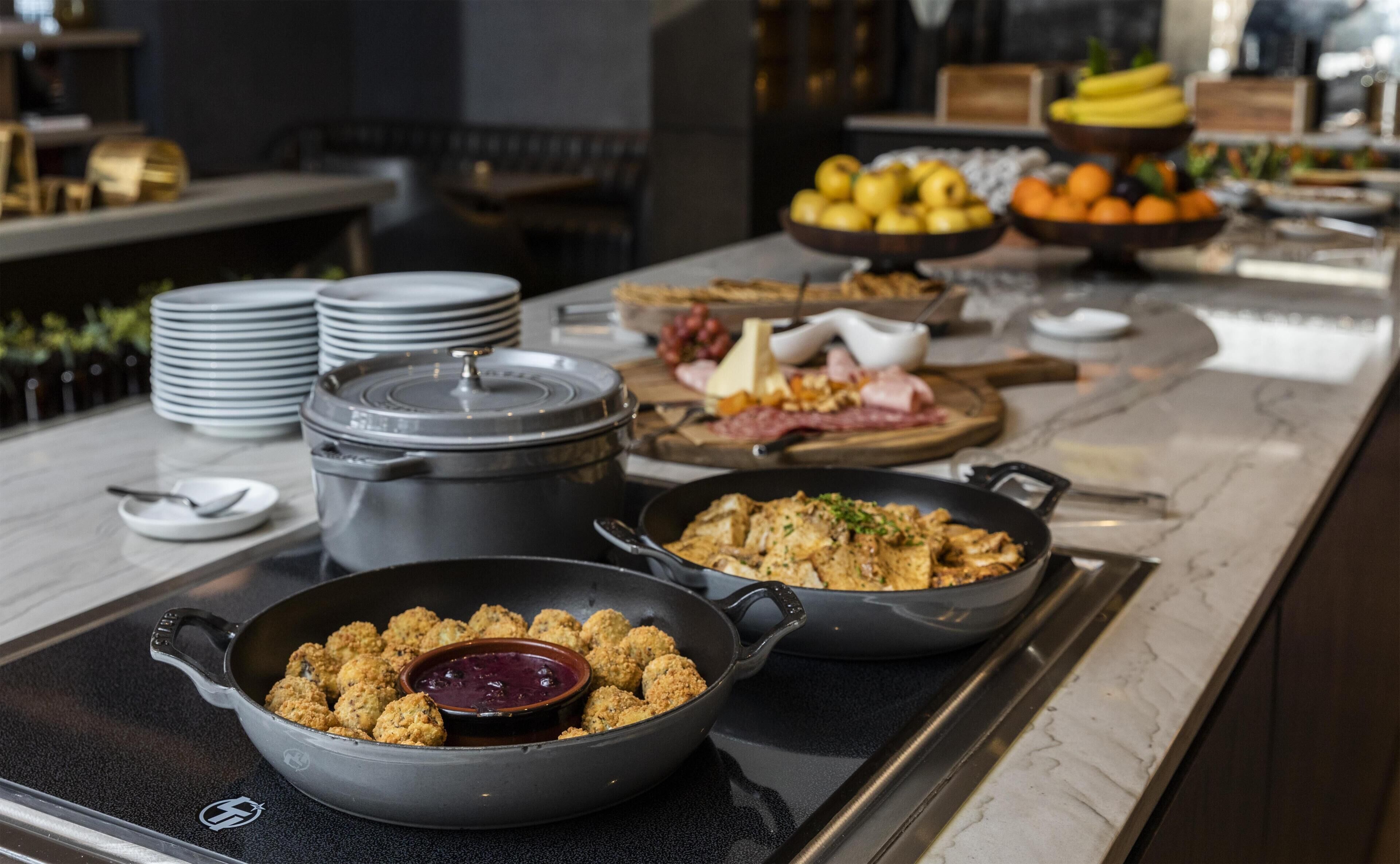 Ein Buffet mit verschiedenen Speisen, darunter eine Schüssel mit Croutons und einer Dipsoße, eine Pfanne mit Rührei und eine Obstauslage im Hintergrund.