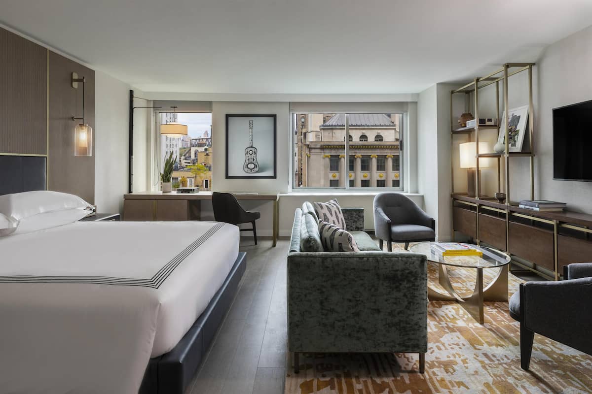 Quarto de hotel com cama grande, sofá, escrivaninha, televisão e vista para a paisagem urbana.