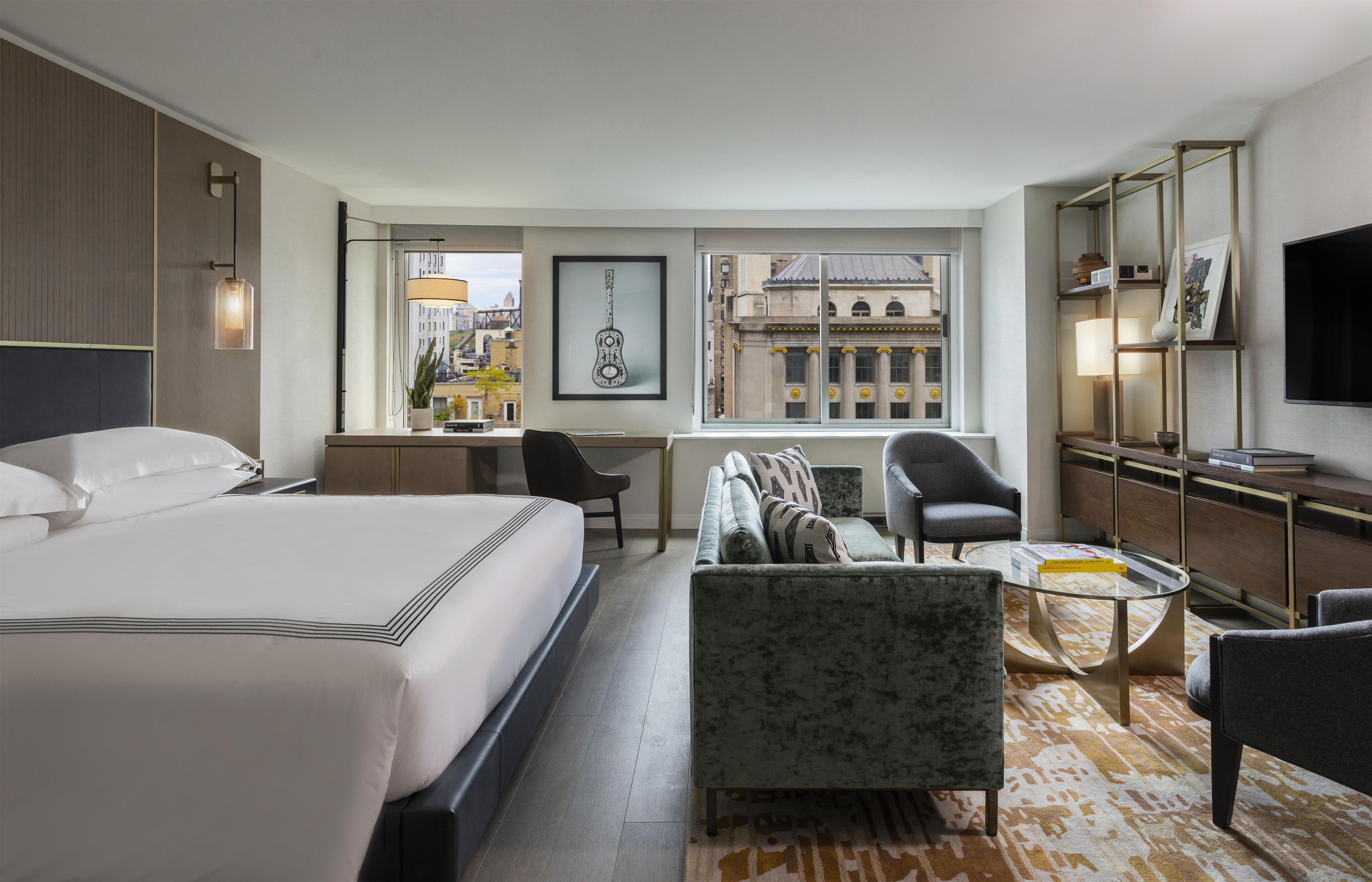 A hotel room with a large bed, a desk, a sofa, a TV, and a view of a cityscape.
