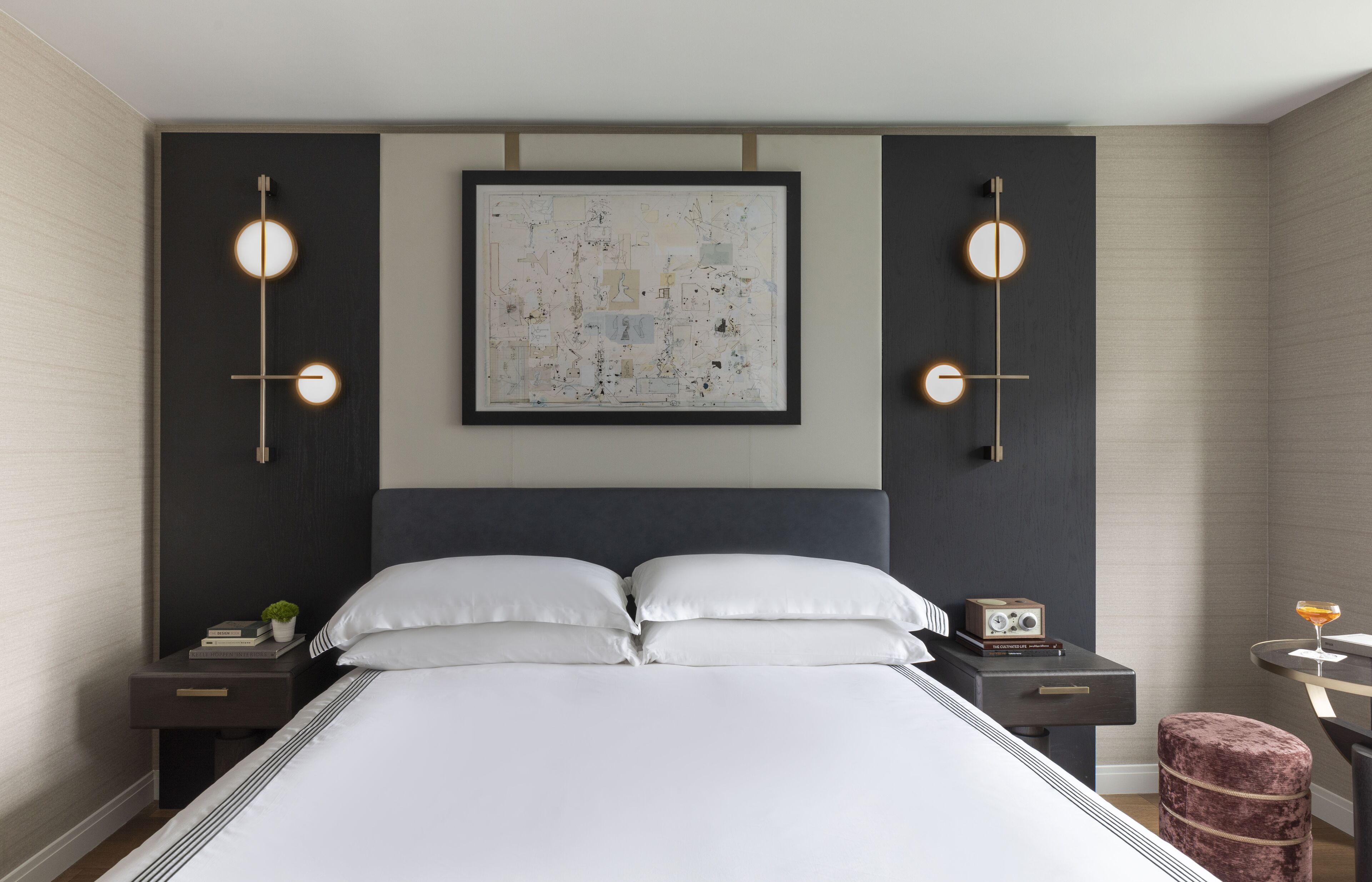 A modern hotel room with a large bed, a framed abstract painting, bedside tables with lamps, and a small round table with a drink.