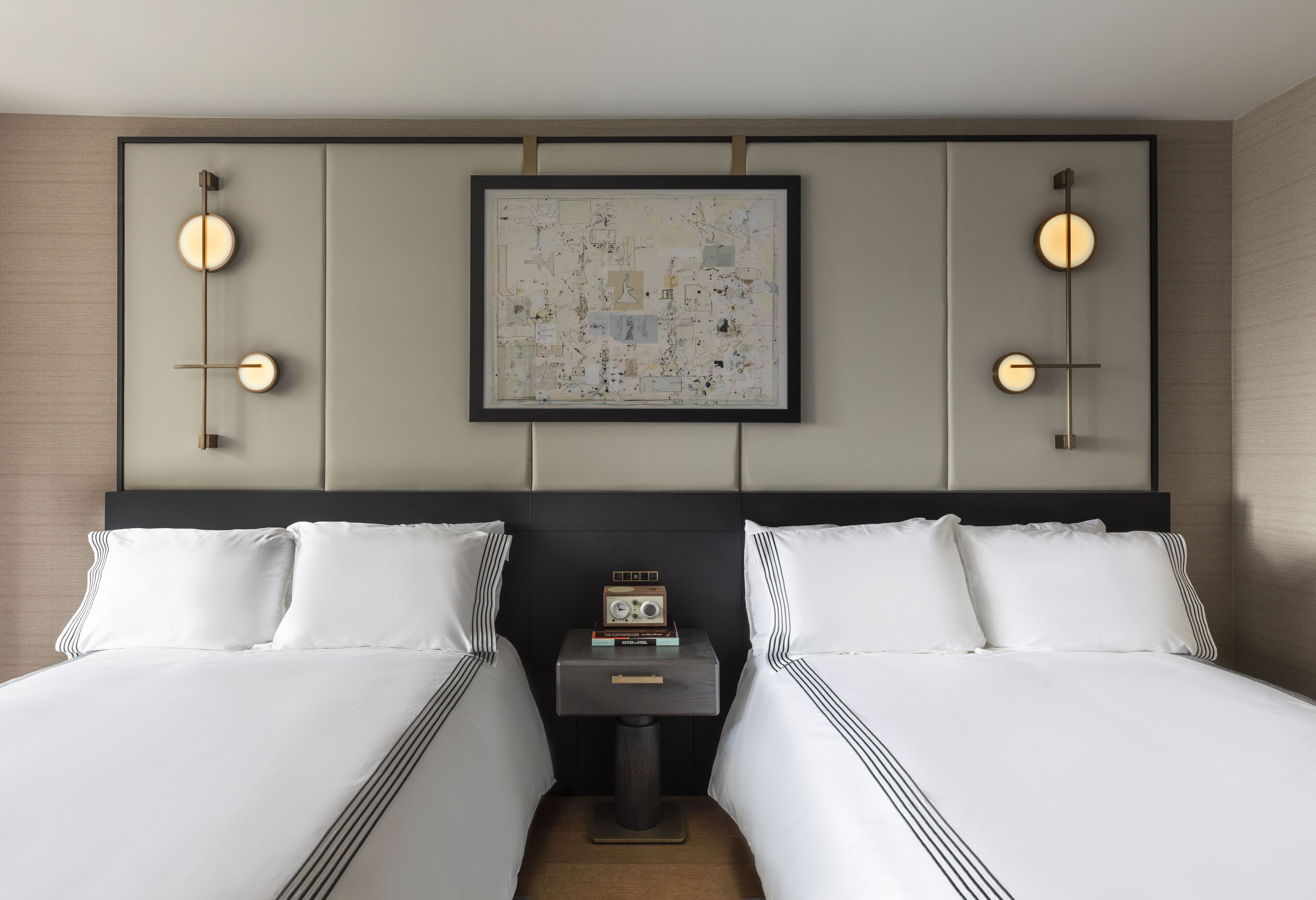 Ein Hotelzimmer mit zwei Betten, einem Nachttisch mit Radio und einem gerahmten abstrakten Gemälde an der Wand.