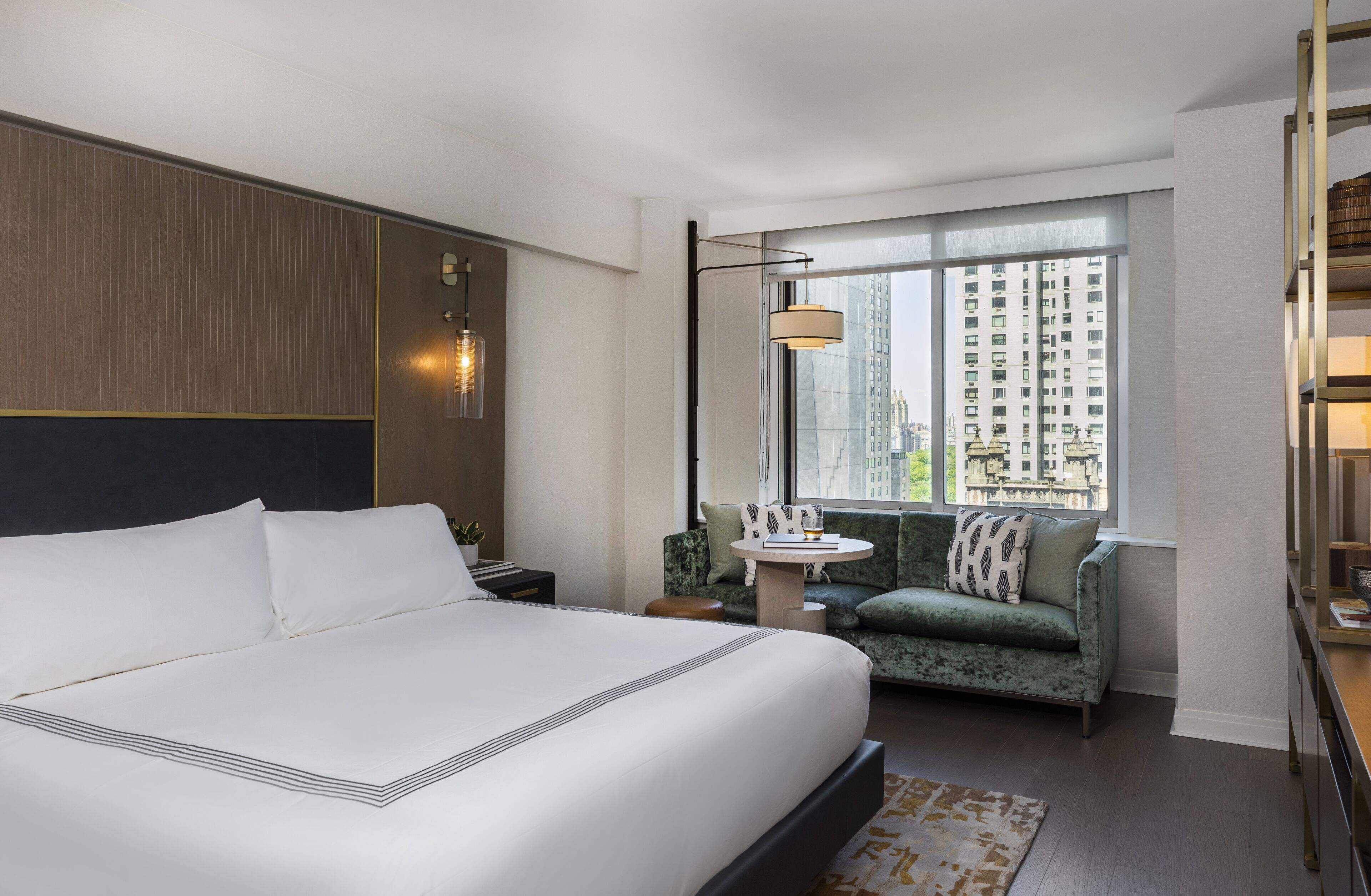 Habitación de hotel moderna con una cama grande, un sofá verde, una pequeña mesa y vistas de edificios altos a través de un amplio ventanal.