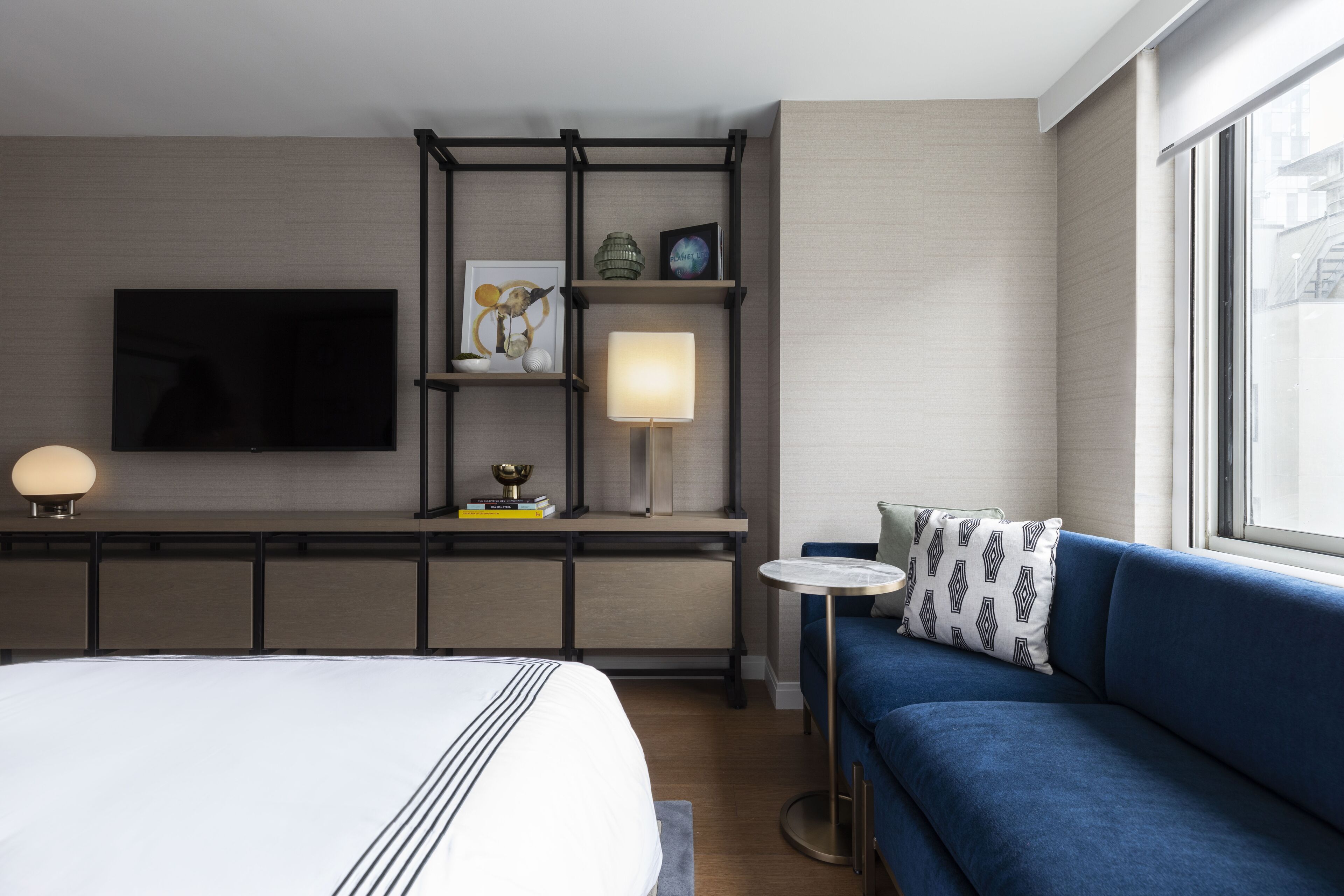 Habitación de hotel con cama, sofá azul, televisor montado en la pared, lámpara y estante con objetos decorativos.
