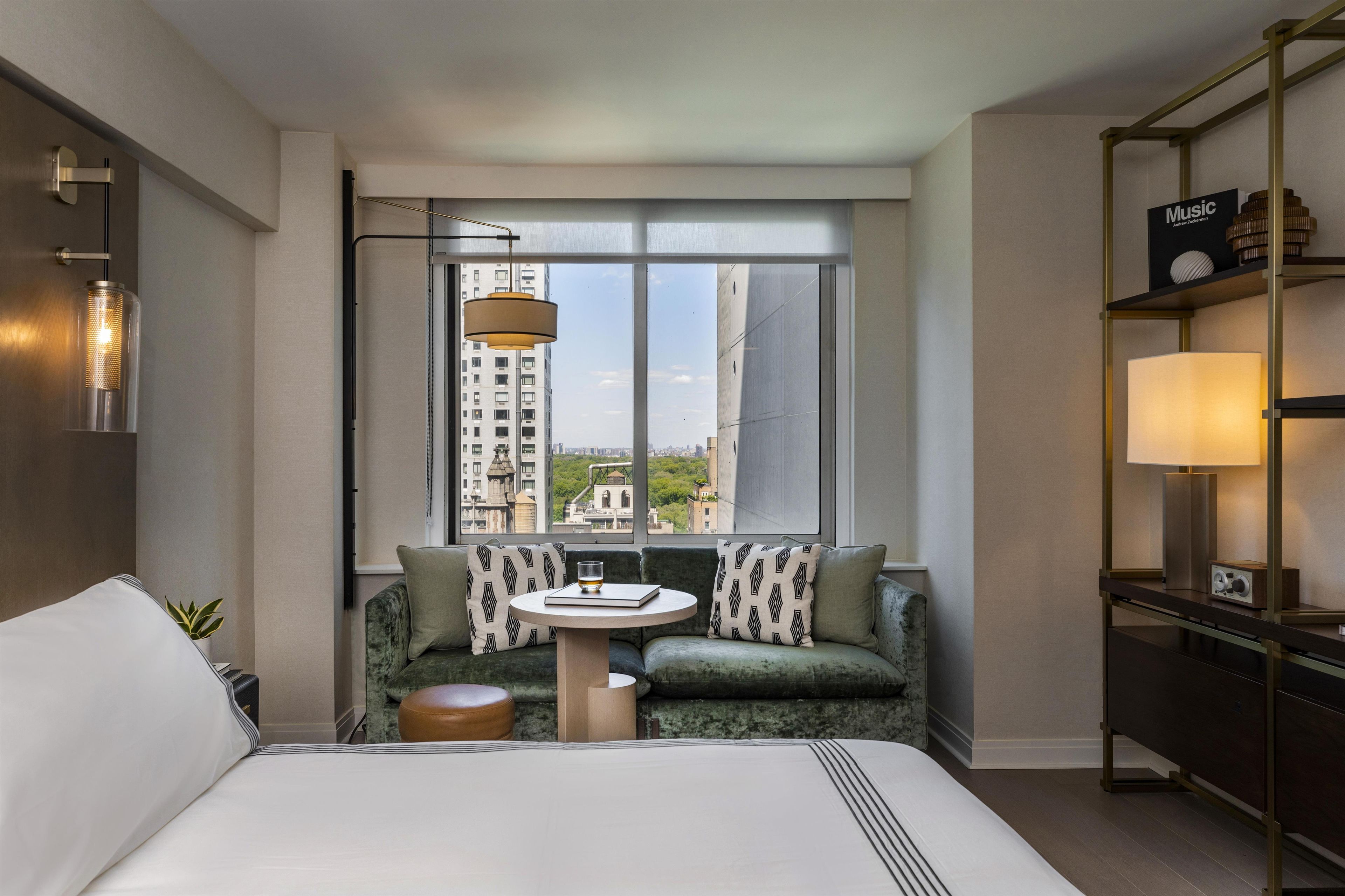 A hotel room with a large bed, a round table with a glass, a sofa with patterned cushions, a bookshelf with a music-themed sign, and a view of a cityscape through a large window.
