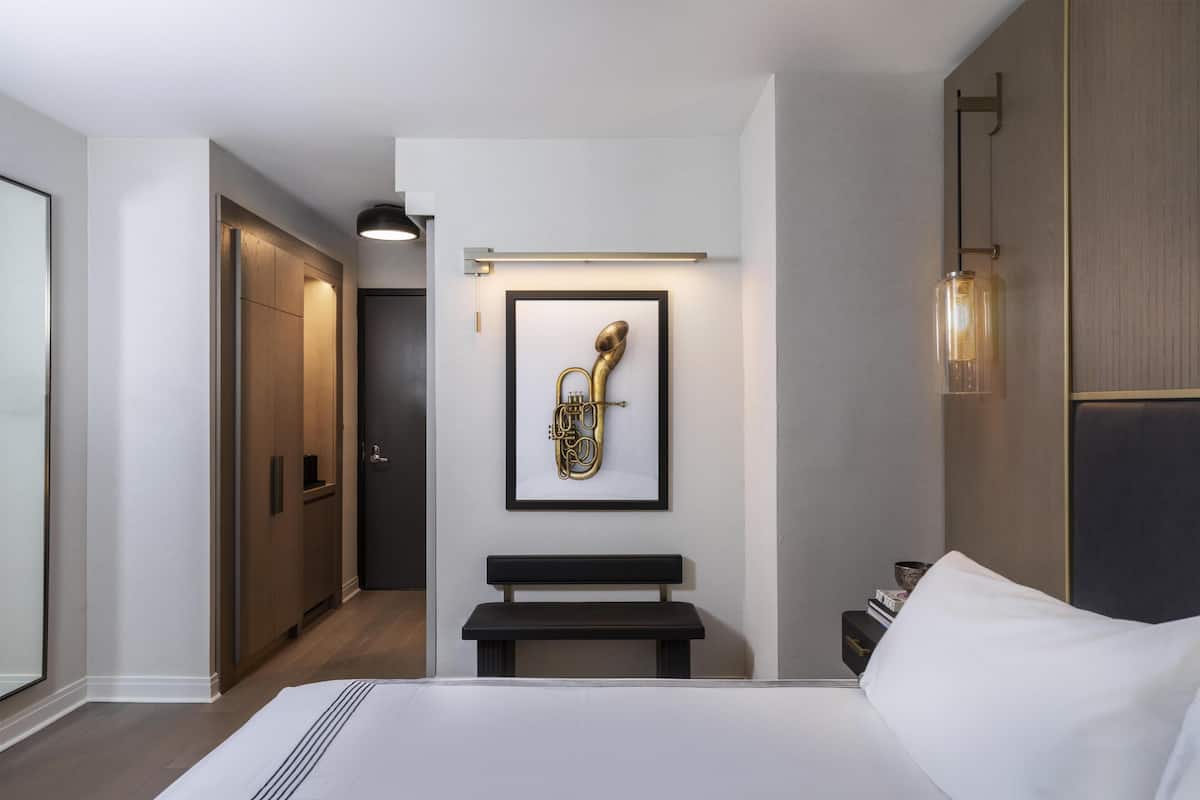 Quarto de hotel com cama, um banco, quadro de um instrumento musical, mesa de cabeceira e guarda-roupa.