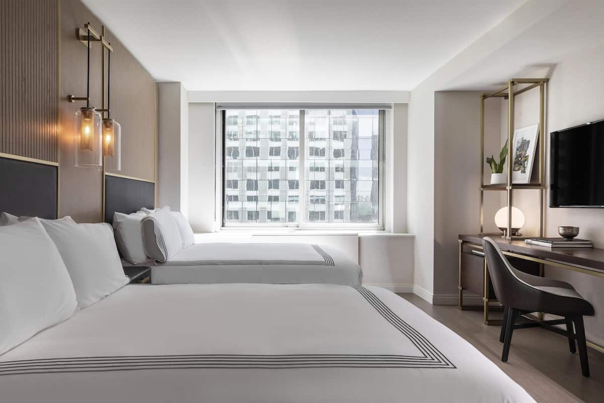 Quarto de hotel com duas camas, uma escrivaninha com cadeira, televisão e uma janela grande com vista para a cidade.