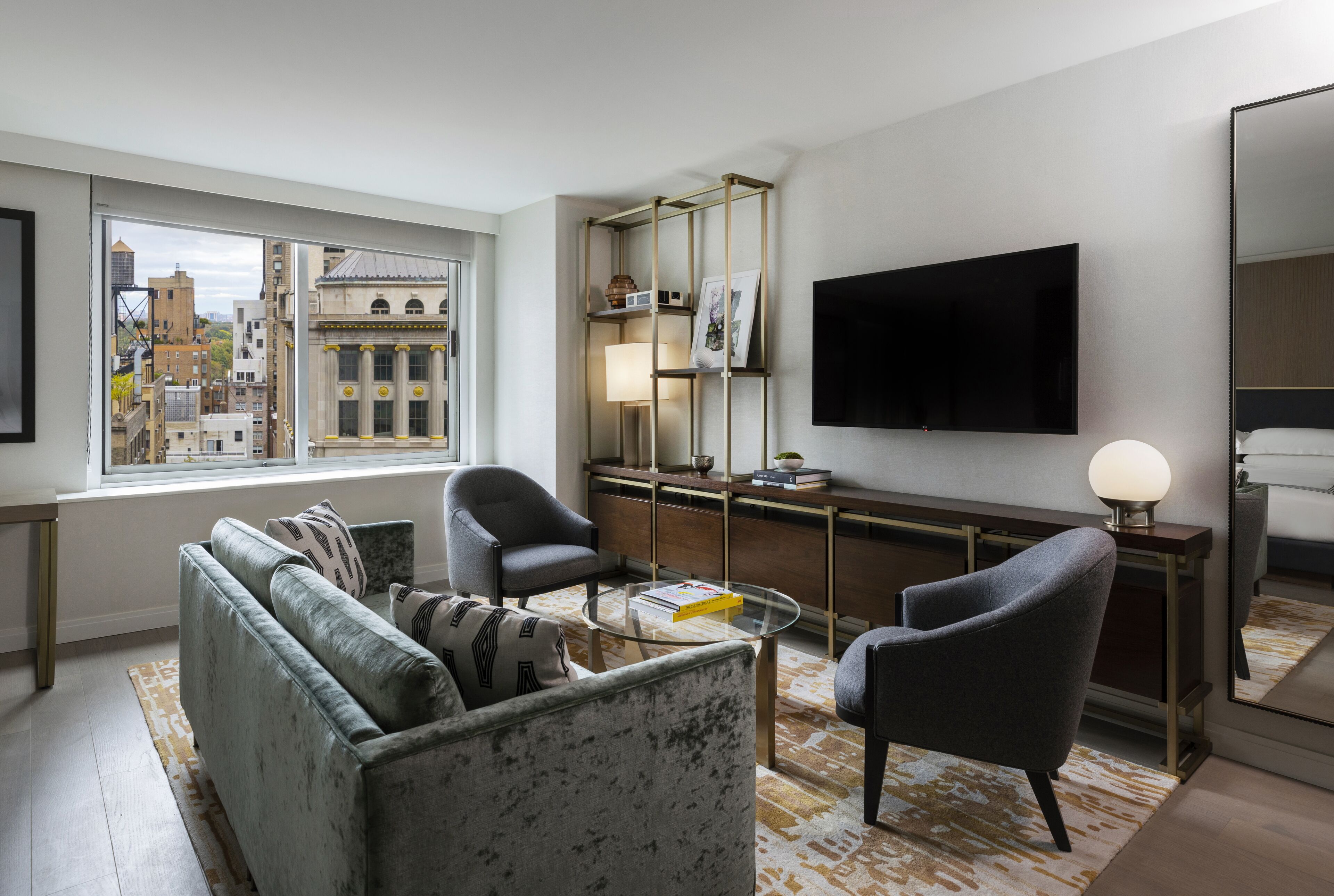 A modern living room with a sofa, armchairs, a coffee table, a TV, and a large window offering a city view.