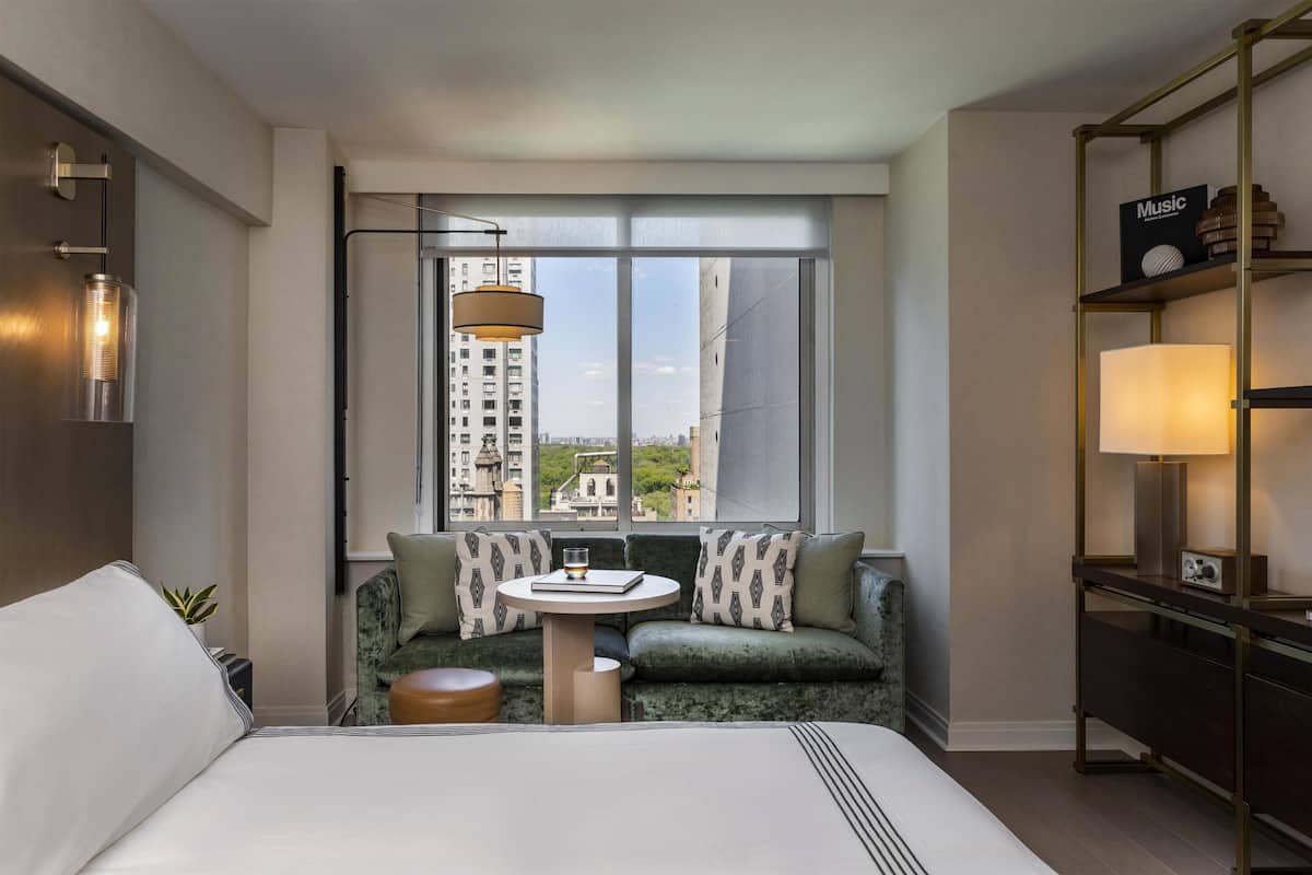 Quarto de hotel com cama, mesa redonda, sofá com almofadas estampadas, estante de livros e uma janela grande com vista para a cidade.