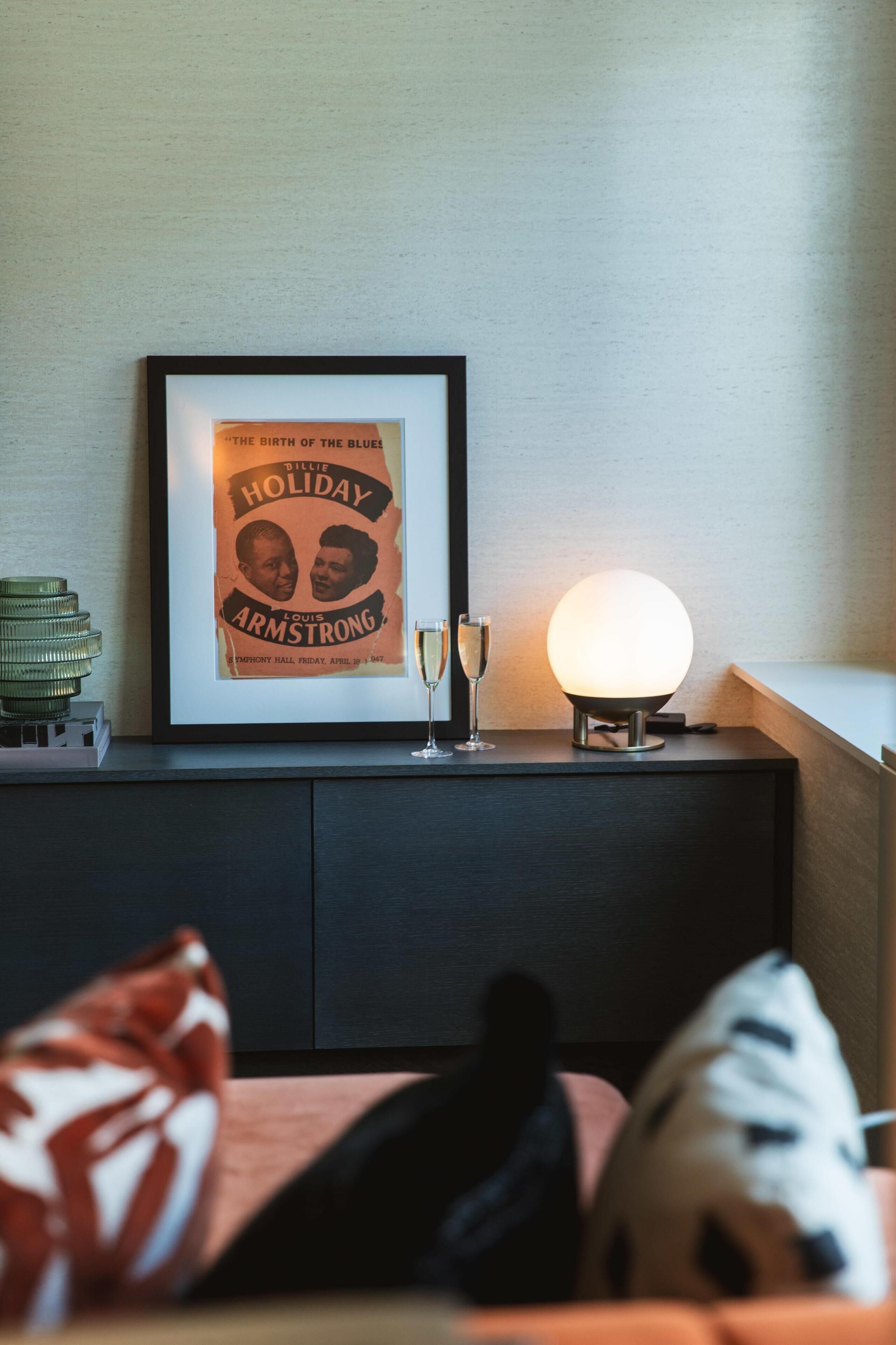 A framed poster on a wall, a lit spherical lamp on a table, and a stack of coins on a side table.