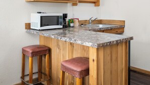 A compact kitchen with wooden cabinets, a built-in microwave, a sink, and two stools.