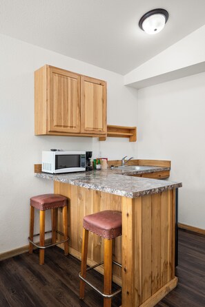 A compact kitchen with wooden cabinets, a built-in microwave, a sink, and two stools.