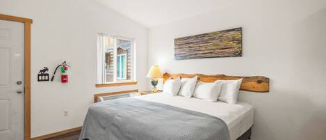 A neatly arranged bedroom with a bed, a window, a painting, and a nightstand with a lamp.