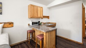 A compact kitchen with wooden cabinets, a microwave, and a sink. There are two stools and a bed in the adjacent room.