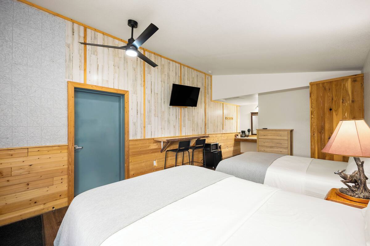 A hotel room with two beds, a desk with chairs, a TV, and a ceiling fan.