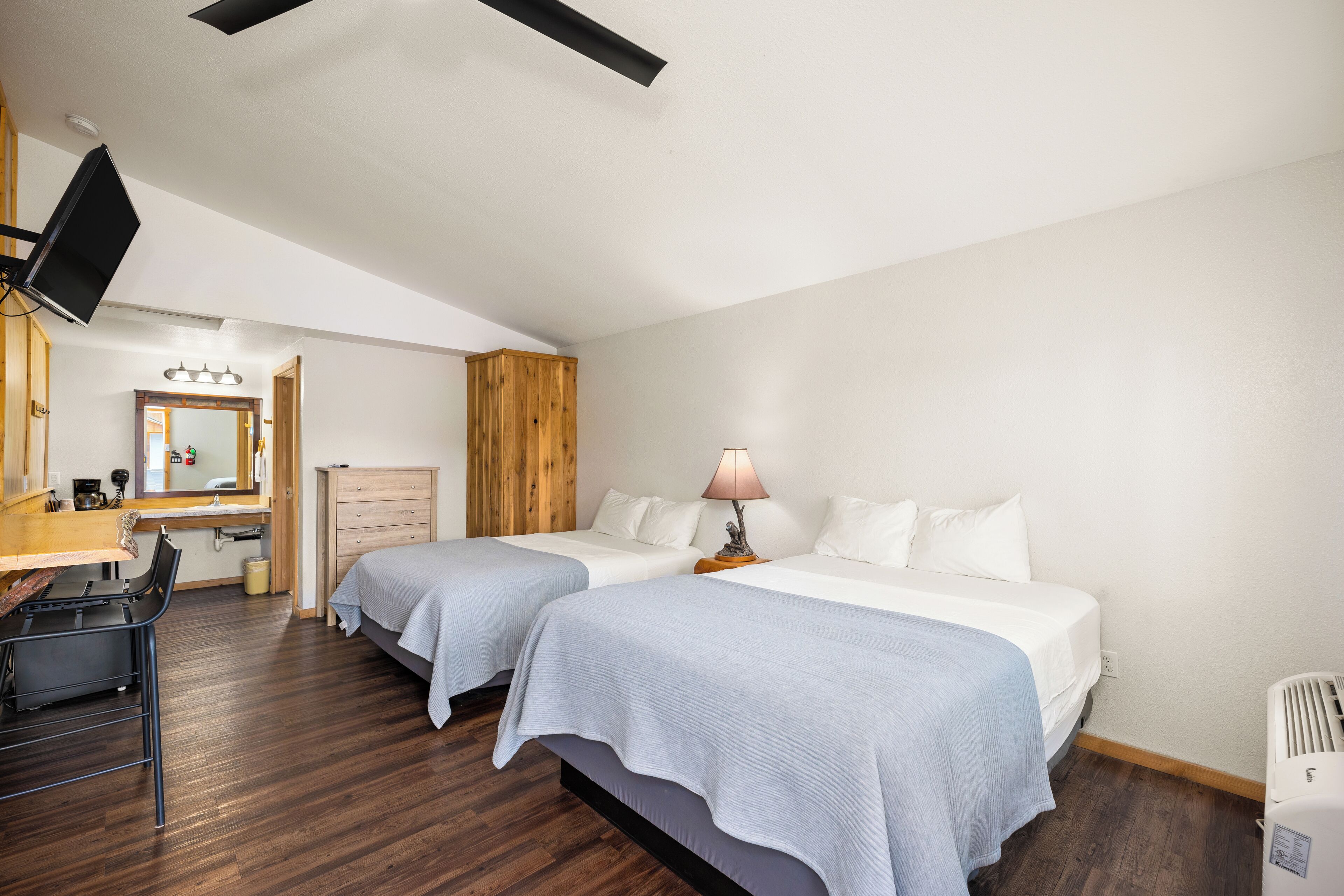 A hotel room with two beds, a desk, a chair, a TV, a mirror, a wooden panel wall, and a ceiling fan.