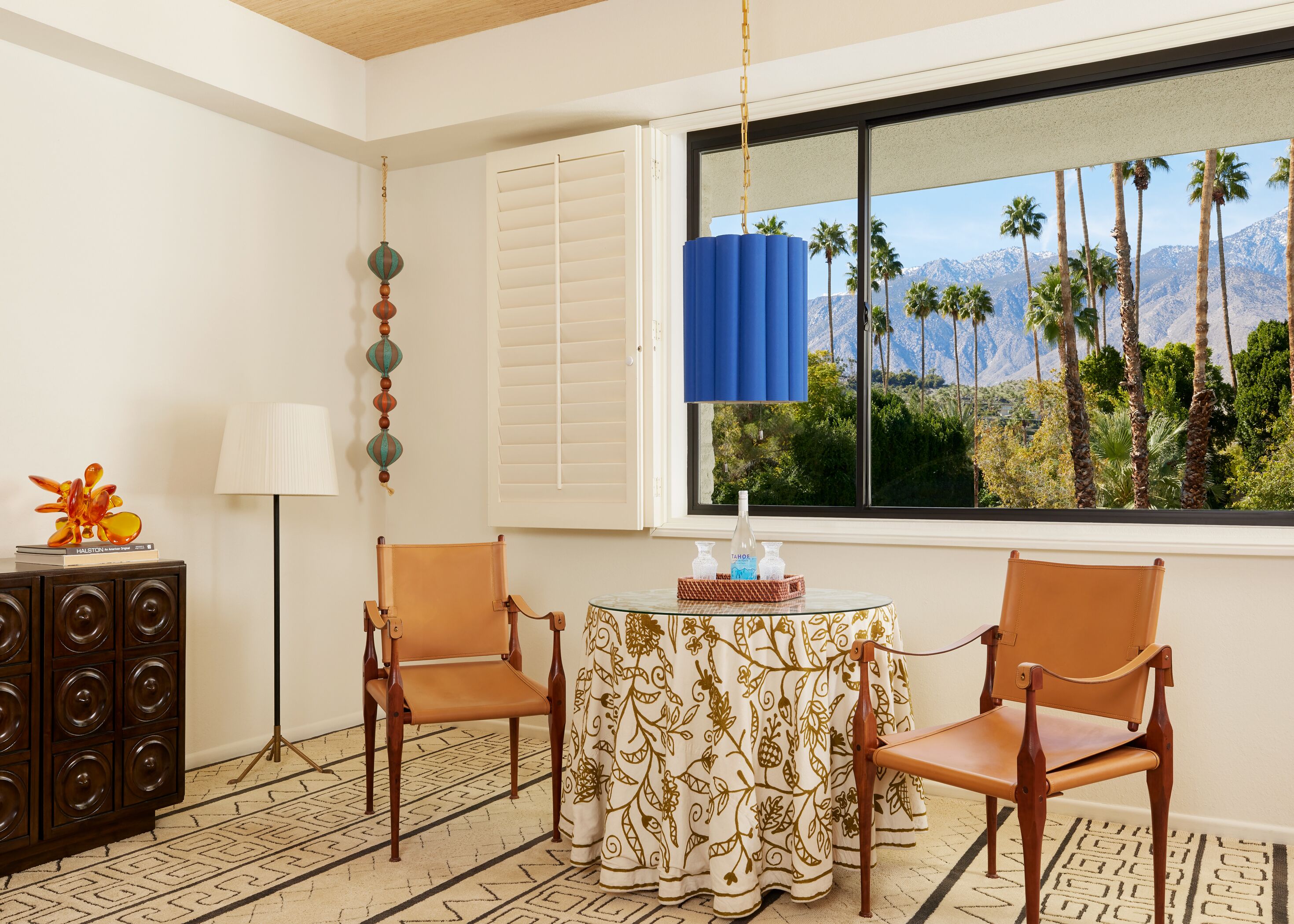 Una habitación con una mesa preparada para dos, un mueble pequeño con altavoces, y vistas a palmeras y montañas a través de una ventana.