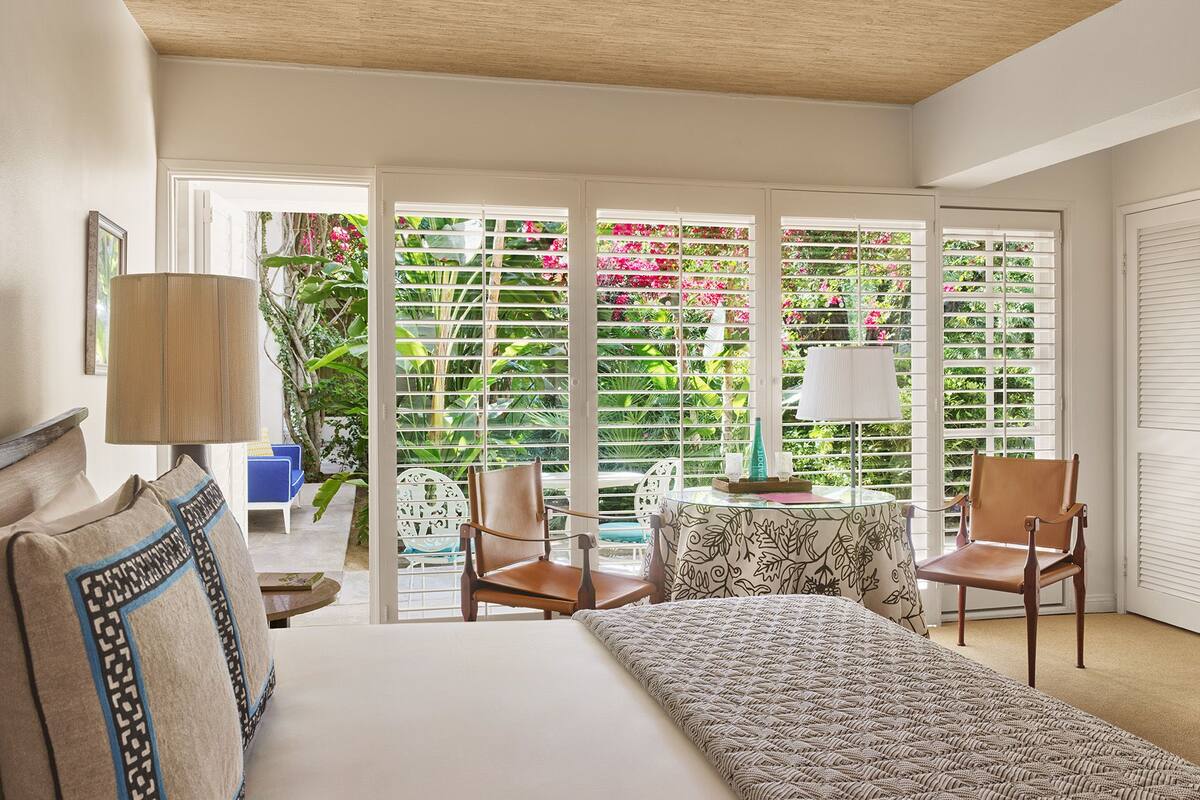 Ein Schlafzimmer mit einem Bett, zwei Stühlen, einem Tisch und Blick auf einen Garten durch große Fenster.