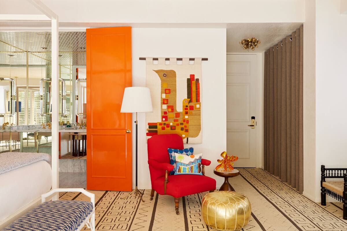 Ein modernes Hotelzimmer mit einer großen orangenen Tür, einem roten Sessel, einem gemusterten Teppich und einem einzigartigen Wandbild.