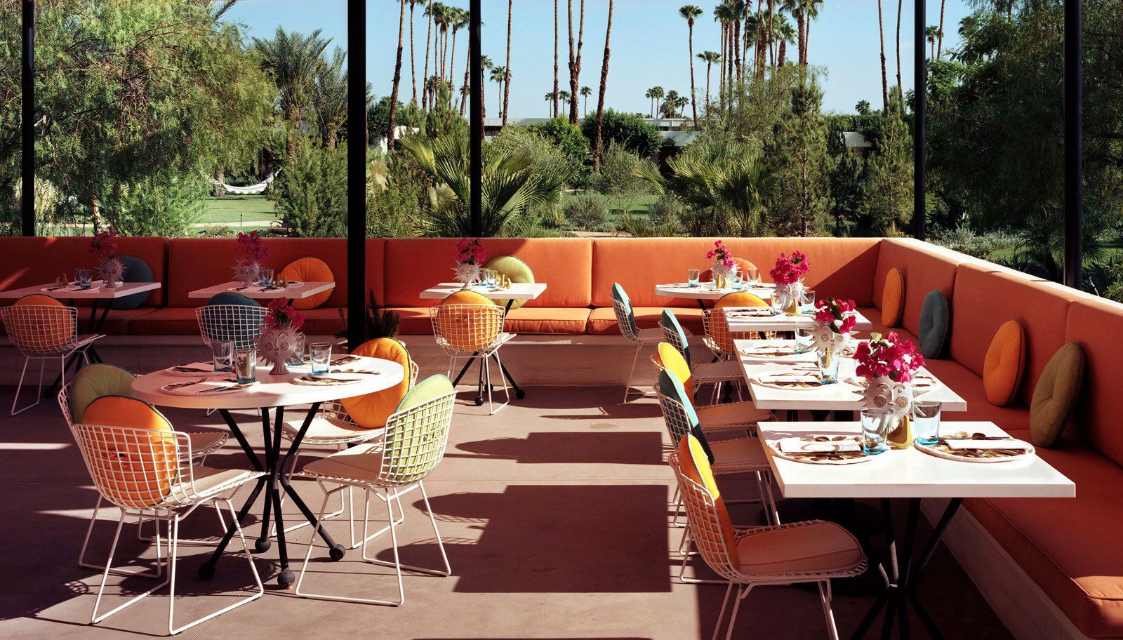 An outdoor dining area with orange seating and white tables, surrounded by greenery and palm trees.