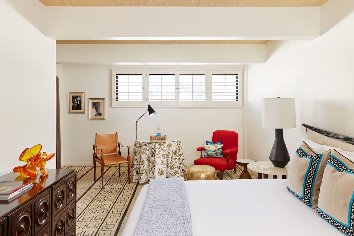 Ein Hotelzimmer mit einem Bett, einem Schreibtisch mit Lampe, einem Sessel, einem kleinen Tisch und einem Fenster mit Jalousien.