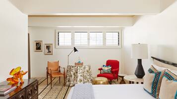 A hotel room with a bed, a desk with a lamp, a chair, a small table, and a window with blinds.
