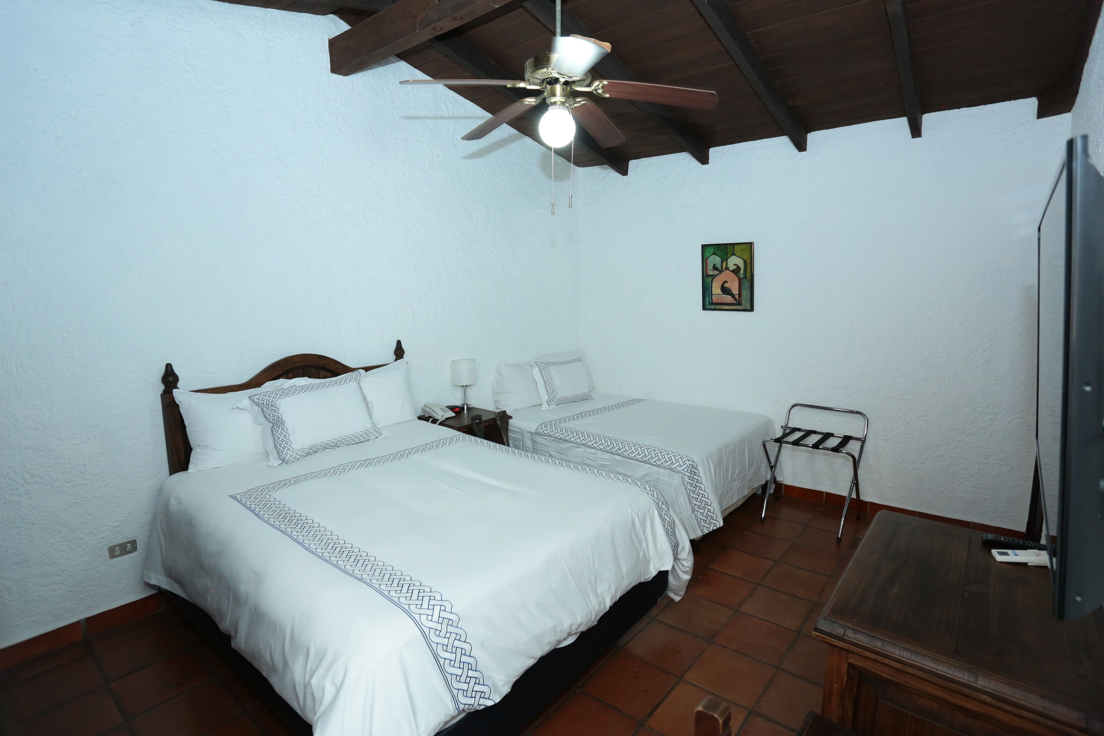Quarto com duas camas, um ventilador de teto, uma mesinha e uma televisão.