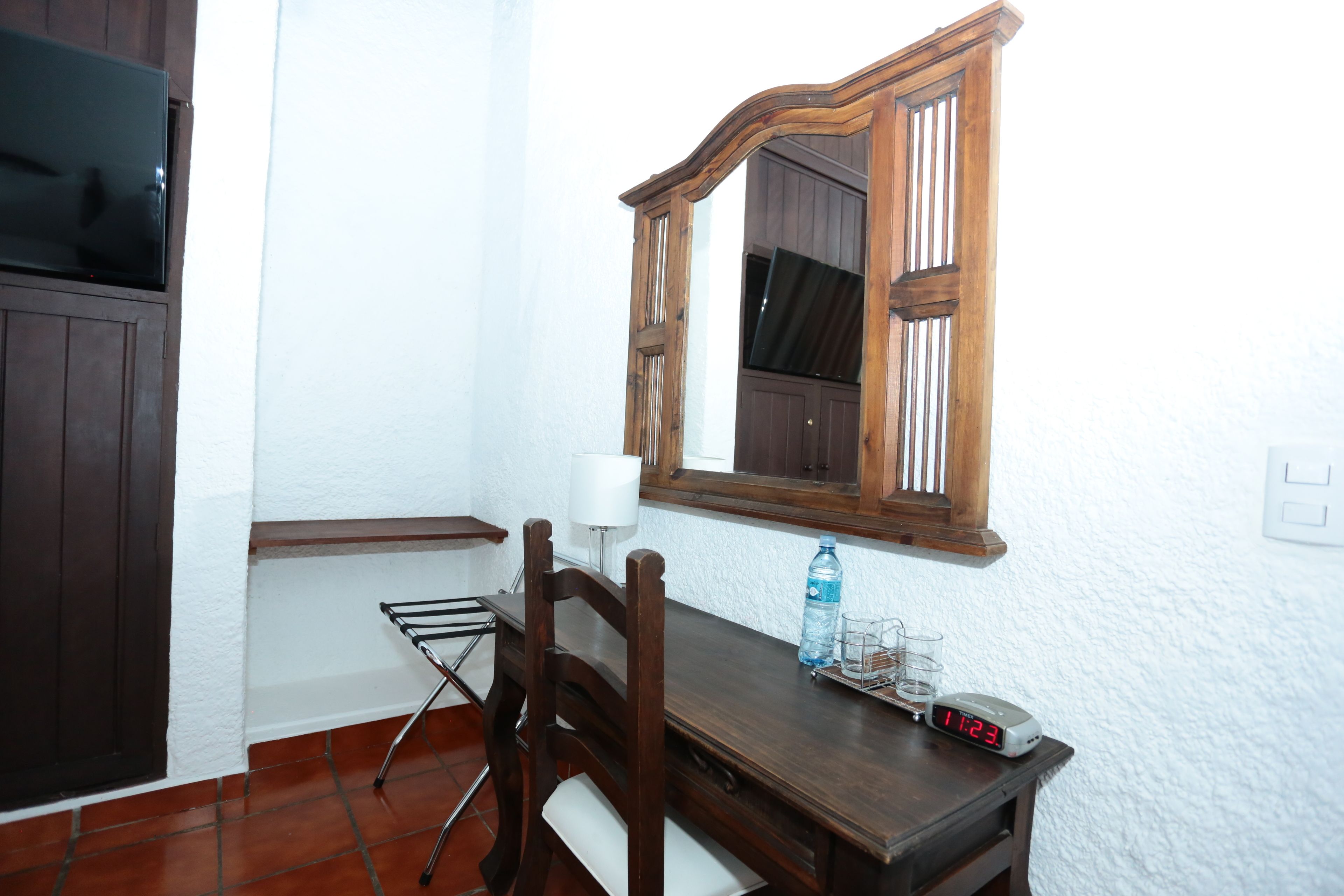 A small, clean room with a wooden table, a chair, a mirror, a clock, and a bottle of water.