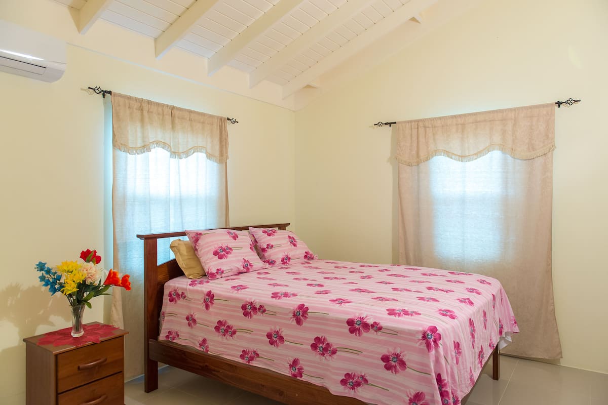 A bedroom with a bed, a nightstand, a vase with flowers, and two windows with curtains.