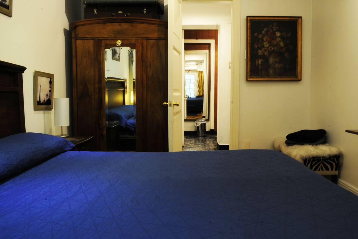 Een hotelkamer met een bed, een kledingkast, een deur naar een andere kamer, een schilderij aan de muur en een klein krukje.