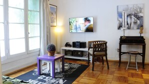 A living room with a television, a piano, a wooden chair, a small table with a plant, and a window with curtains.